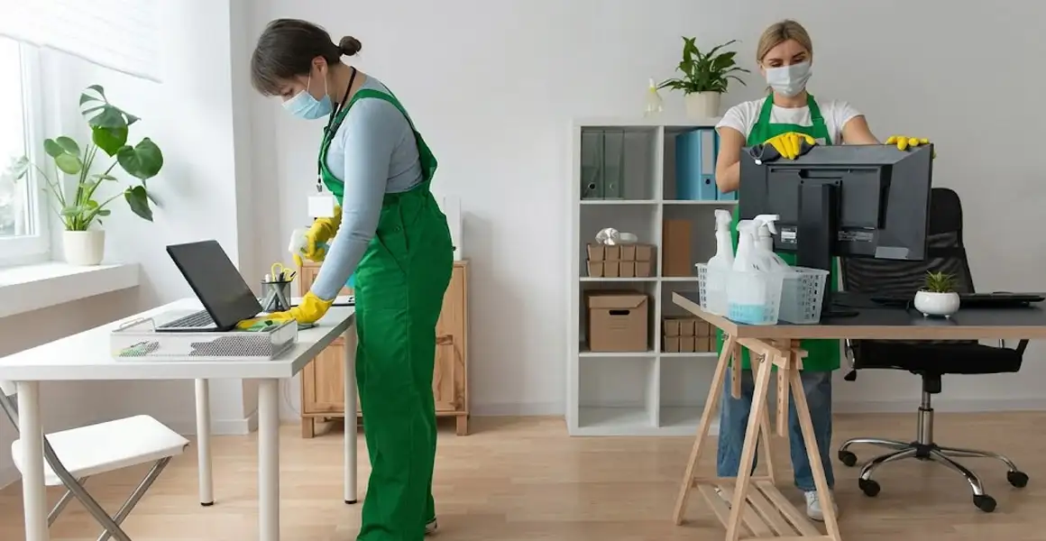 Two professional female cleaners in green uniforms sanitizing desks, laptops, and computer monitors in a Riyadh office. Thorough office cleaning and disinfection services in Riyadh.