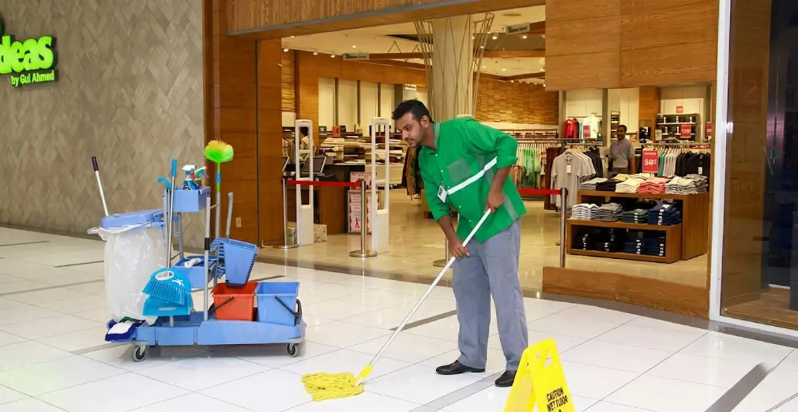 Professional janitorial staff in green uniforms mopping the tiled floor of a shopping mall in Riyadh, featuring a yellow caution wet floor sign and a fully equipped cleaning trolley.