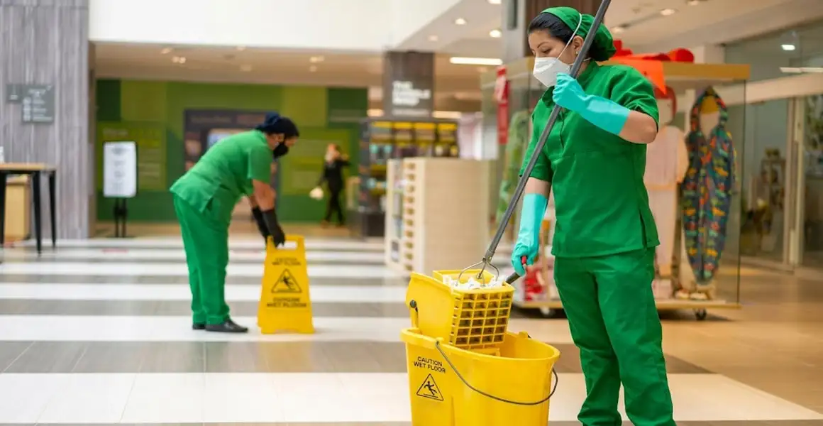 Two commercial cleaning workers wearing face masks and green uniforms sanitizing and mopping a retail mall hallway in Riyadh next to a bright yellow wet floor sign.